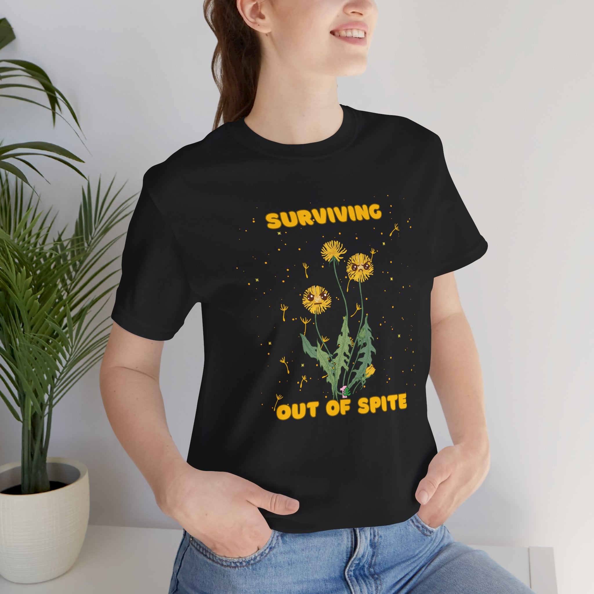 A person wearing a black plant t-shirt with yellow "surviving out of spite" funny text and dandelion plants with a funny look printed on the front of the shirt.