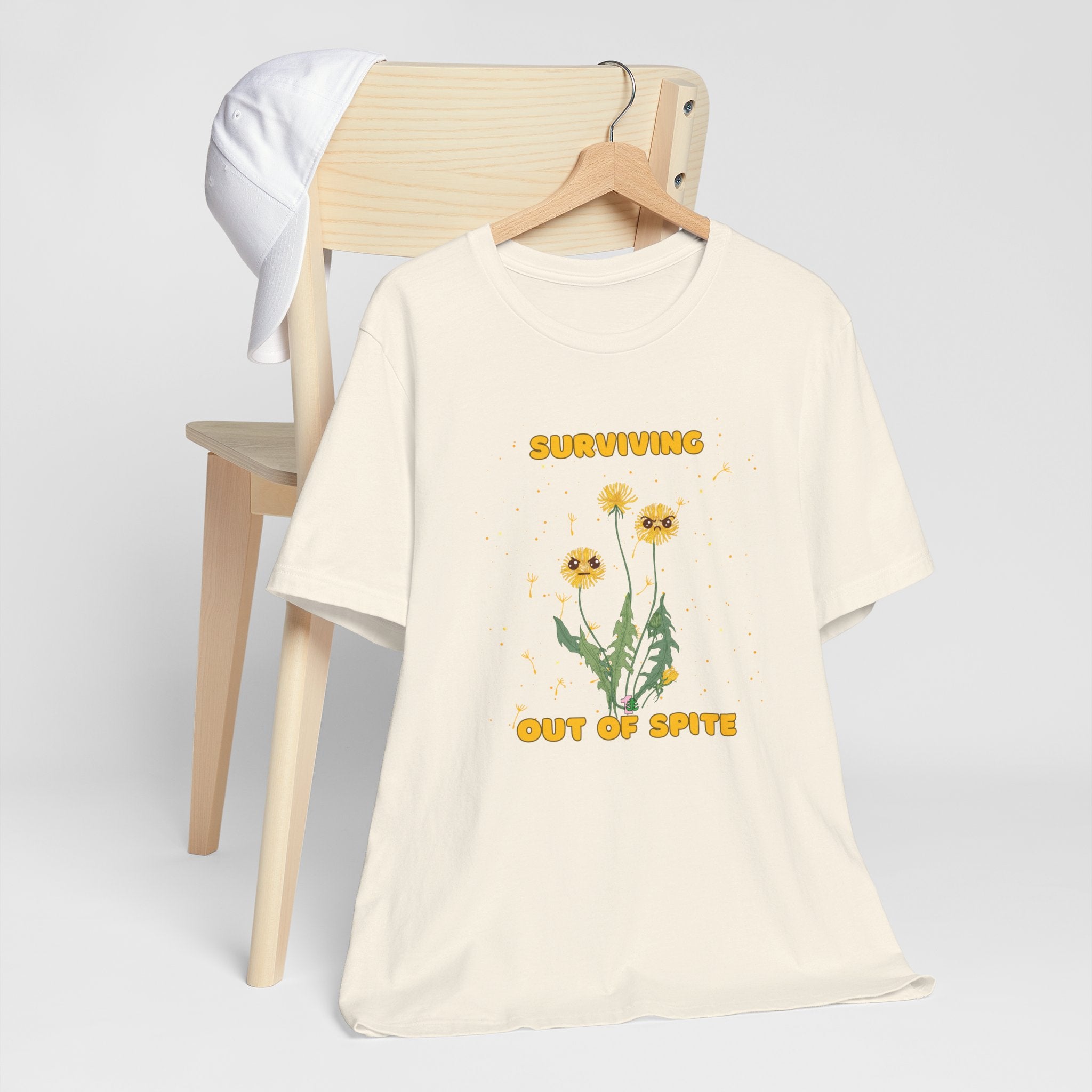 A hanging natural color t-shirt with yellow "surviving out of spite" funny text and dandelion plants with a funny look printed on the front of the shirt.