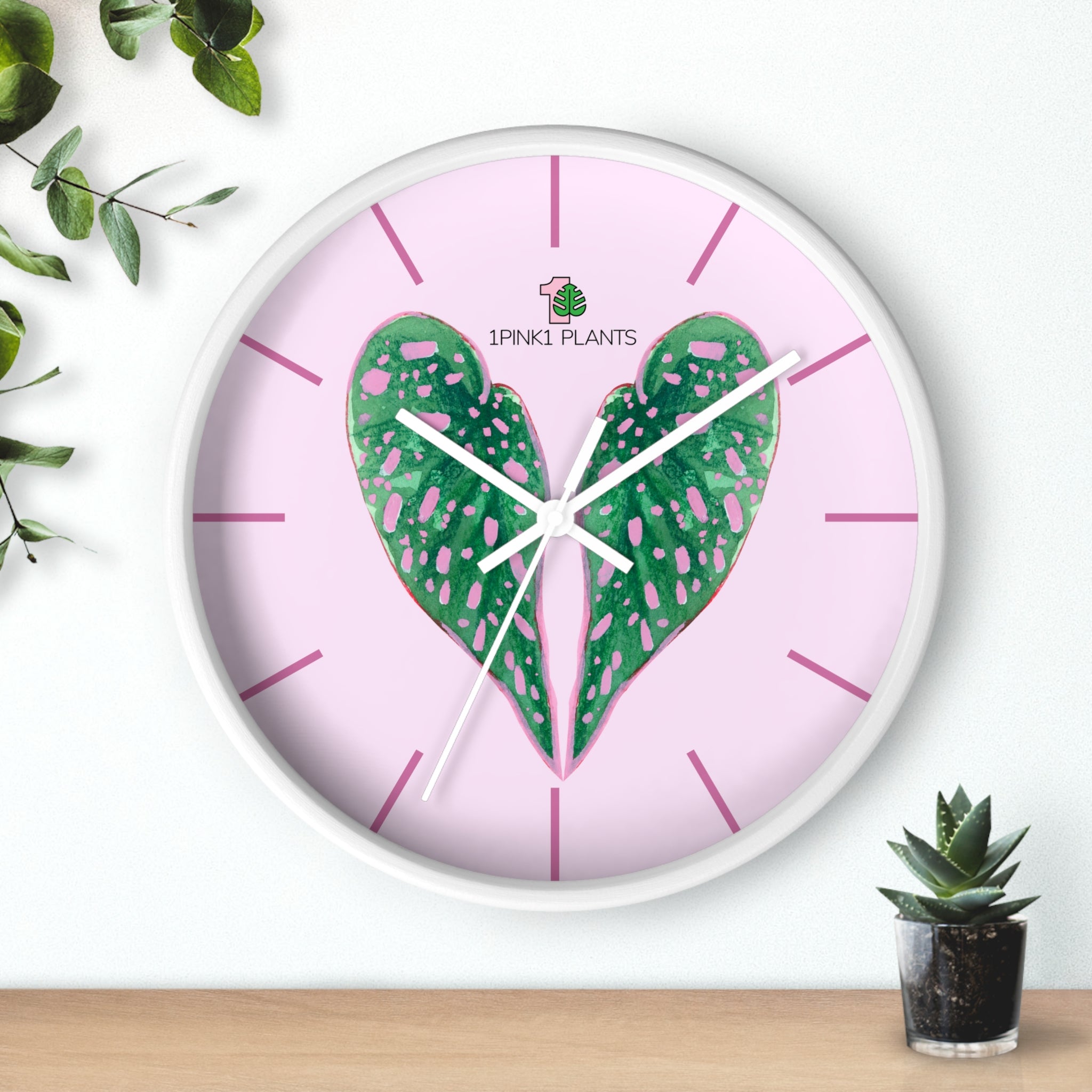 A picture of a circular wall clock.  It has a design of begonia plant leaves in the shape of a heart and a pink background. 