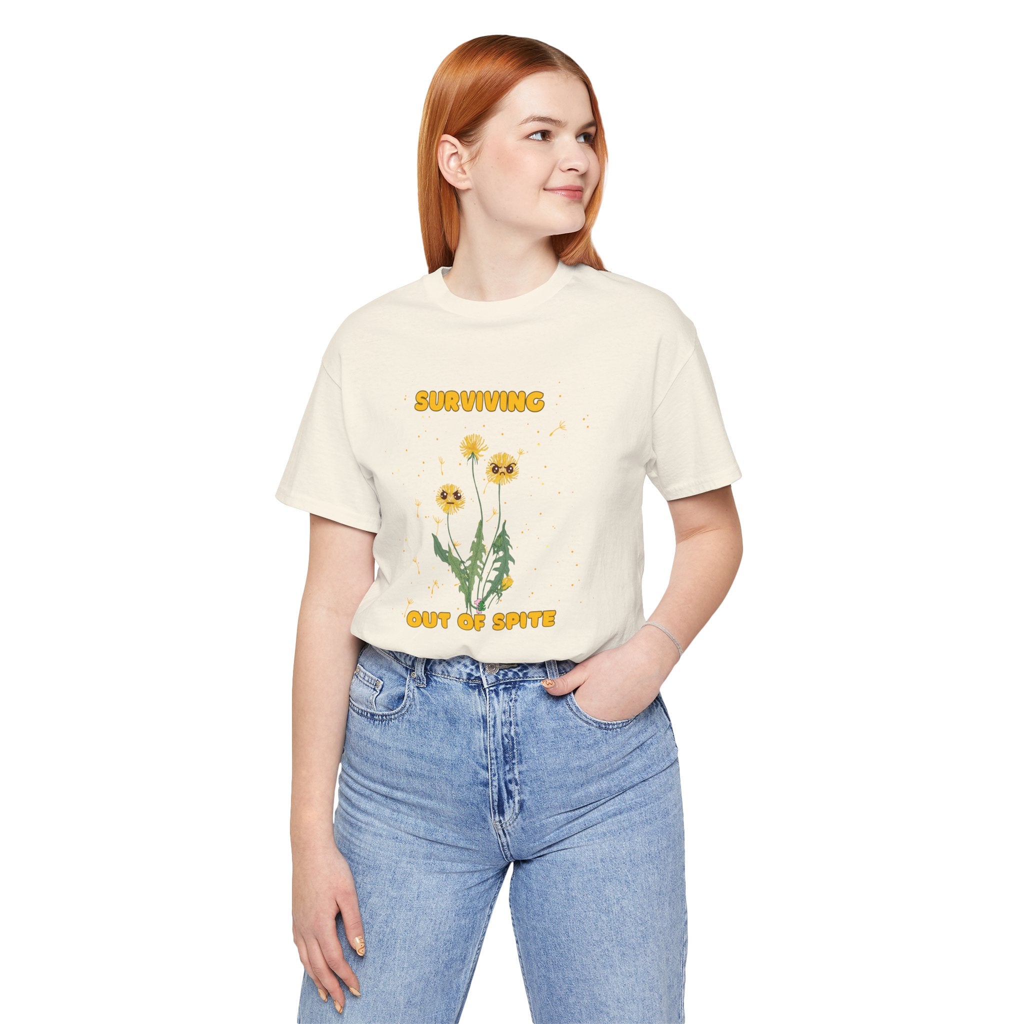 A person wearing a natural color t-shirt with yellow "surviving out of spite" funny text and dandelion plants with a funny look printed on the front of the shirt. 