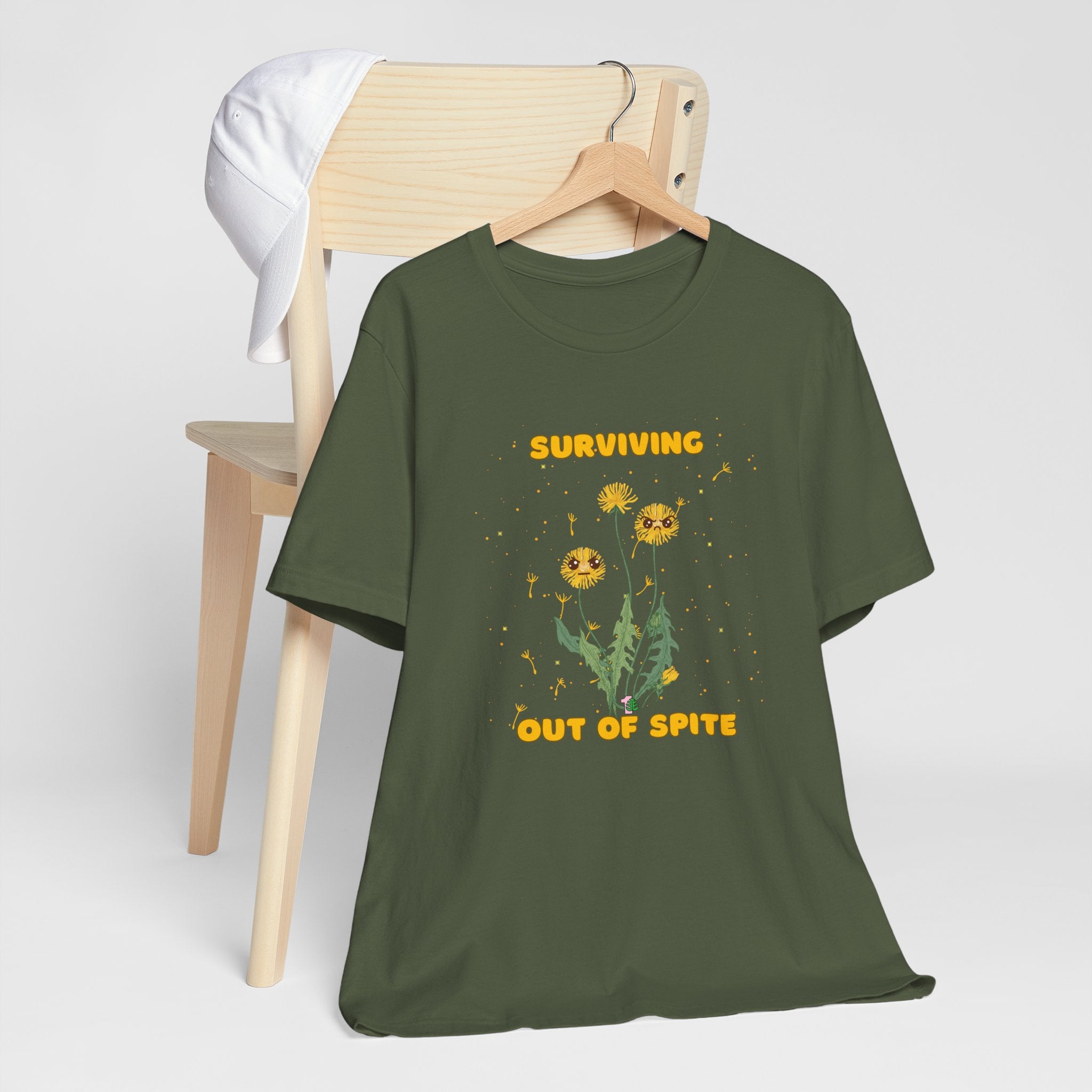 A military green color t-shirt with yellow "surviving out of spite" funny text.  There is also dandelion plants with a funny look printed on the front of the shirt. 