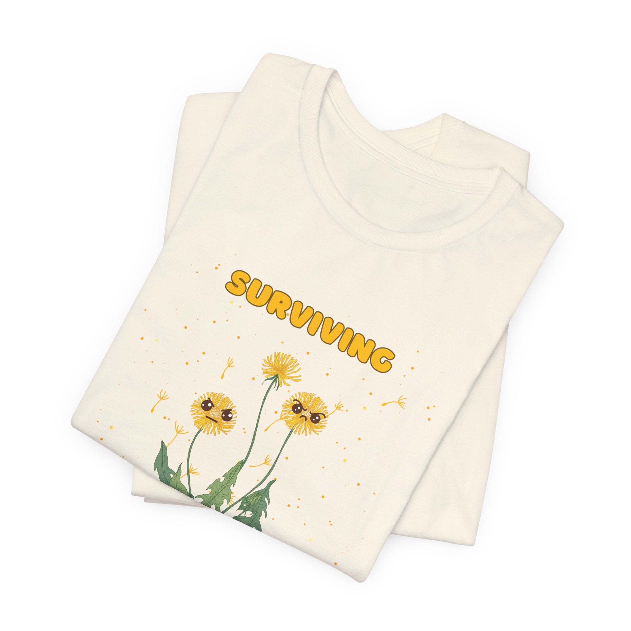 A folded natural color t-shirt with yellow "surviving out of spite" funny text and dandelion plants with a funny look printed on the front of the shirt. 