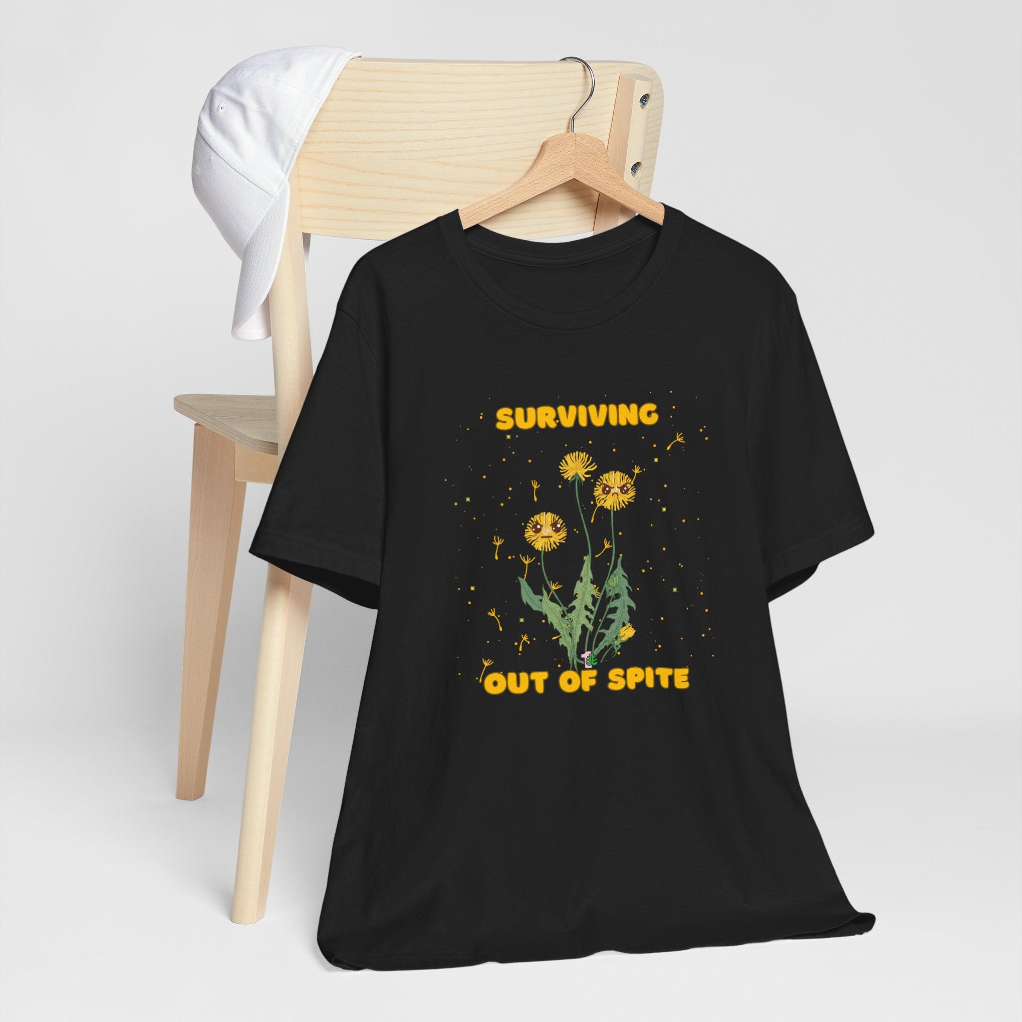 A hanging black color t-shirt with yellow "surviving out of spite" funny text and dandelion plants with a funny look printed on the front of the shirt. 