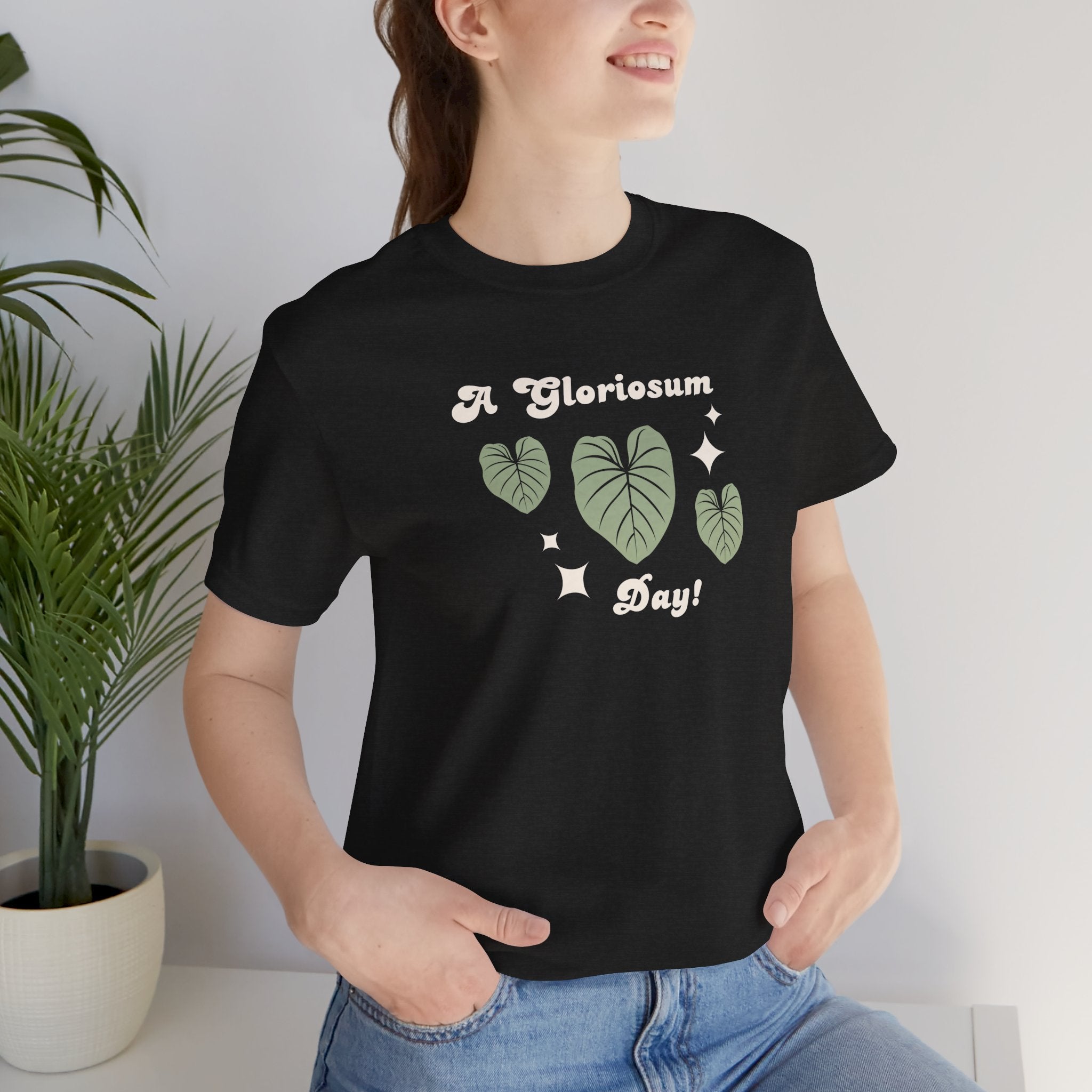 A person wearing a cute plat tshirt with a philodendron gloriosum design and text that says "A gloriosum Day!" An excellent gift for plant lovers, plant collectors, and plant enthusiasts. Great as a birthday gift. This folded t-shirt comes in a black Heather color.