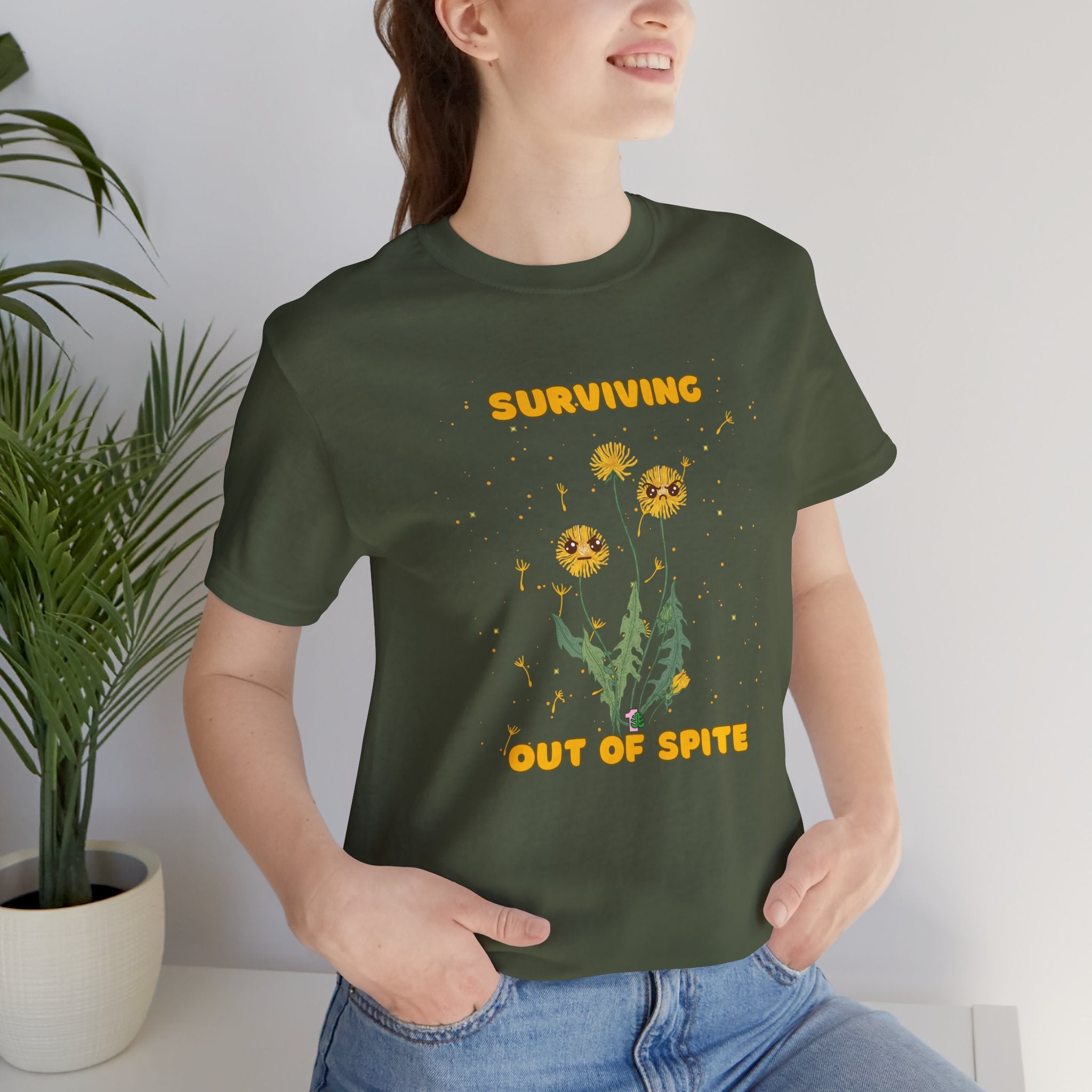 A person wearing a military green color t-shirt with yellow "surviving out of spite" funny text and dandelion plants with a funny look printed on the front of the shirt. 