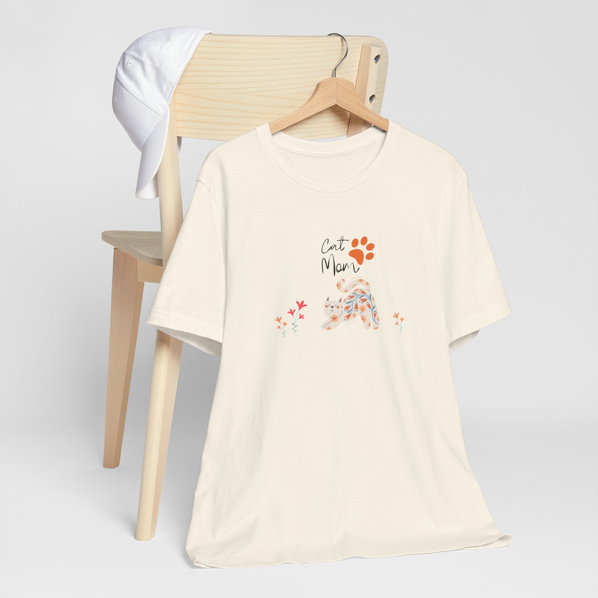 A natural color t-shirt with the words "cat mom," a floral cat, and some flowers printed on the front of the shirt.