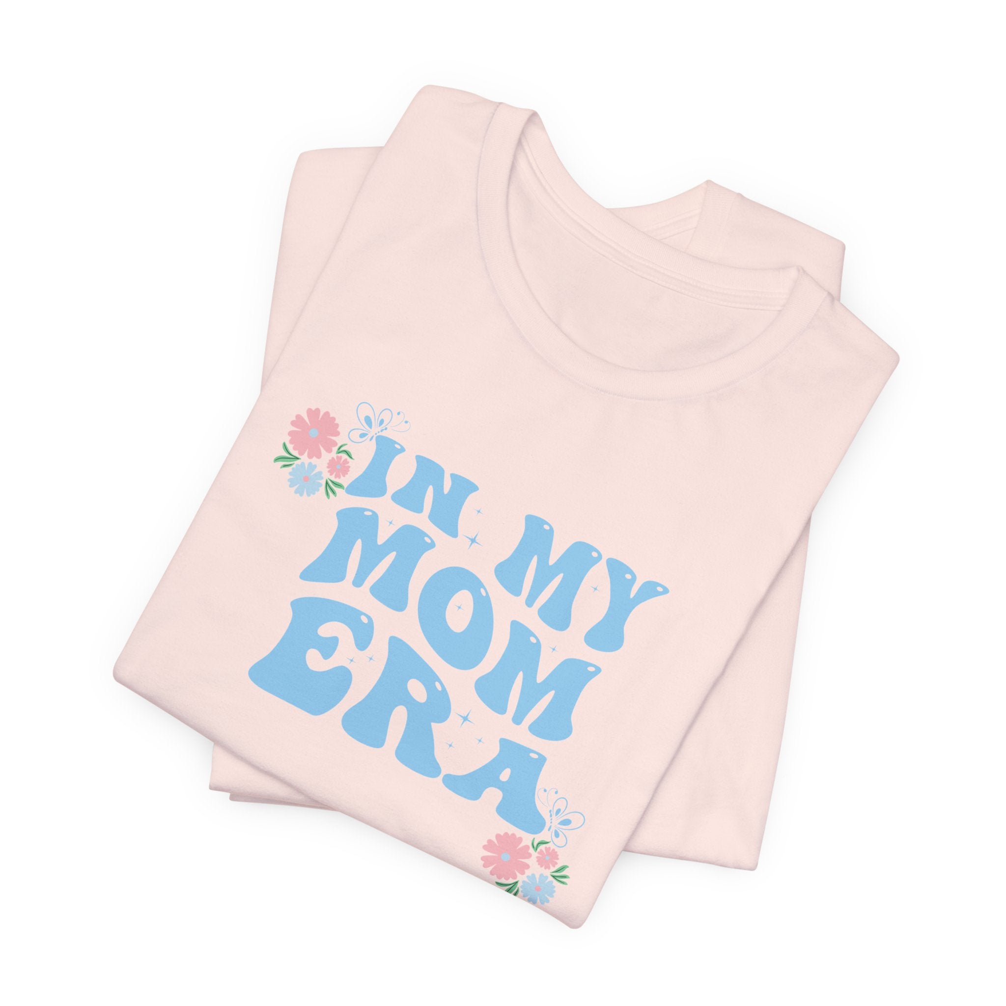 a pink folded tee shirt with the words "In my mom era" printed in blue. This shirt makes an excellent t-shirt for soon to be moms.