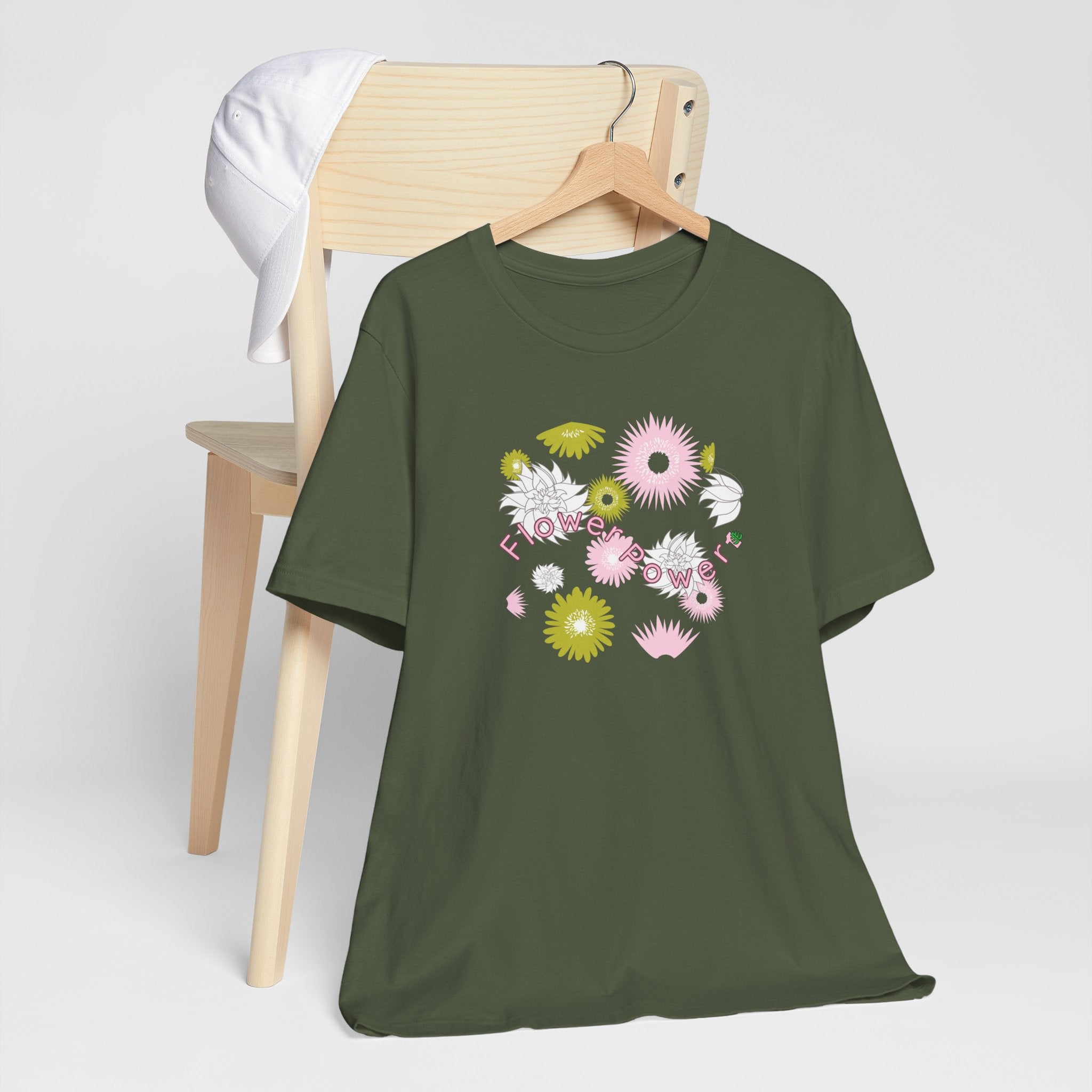 A hanging military green color tee shirt with the words flower power in pink.  There are also pink, white, and green flowers printed on the shirt. 