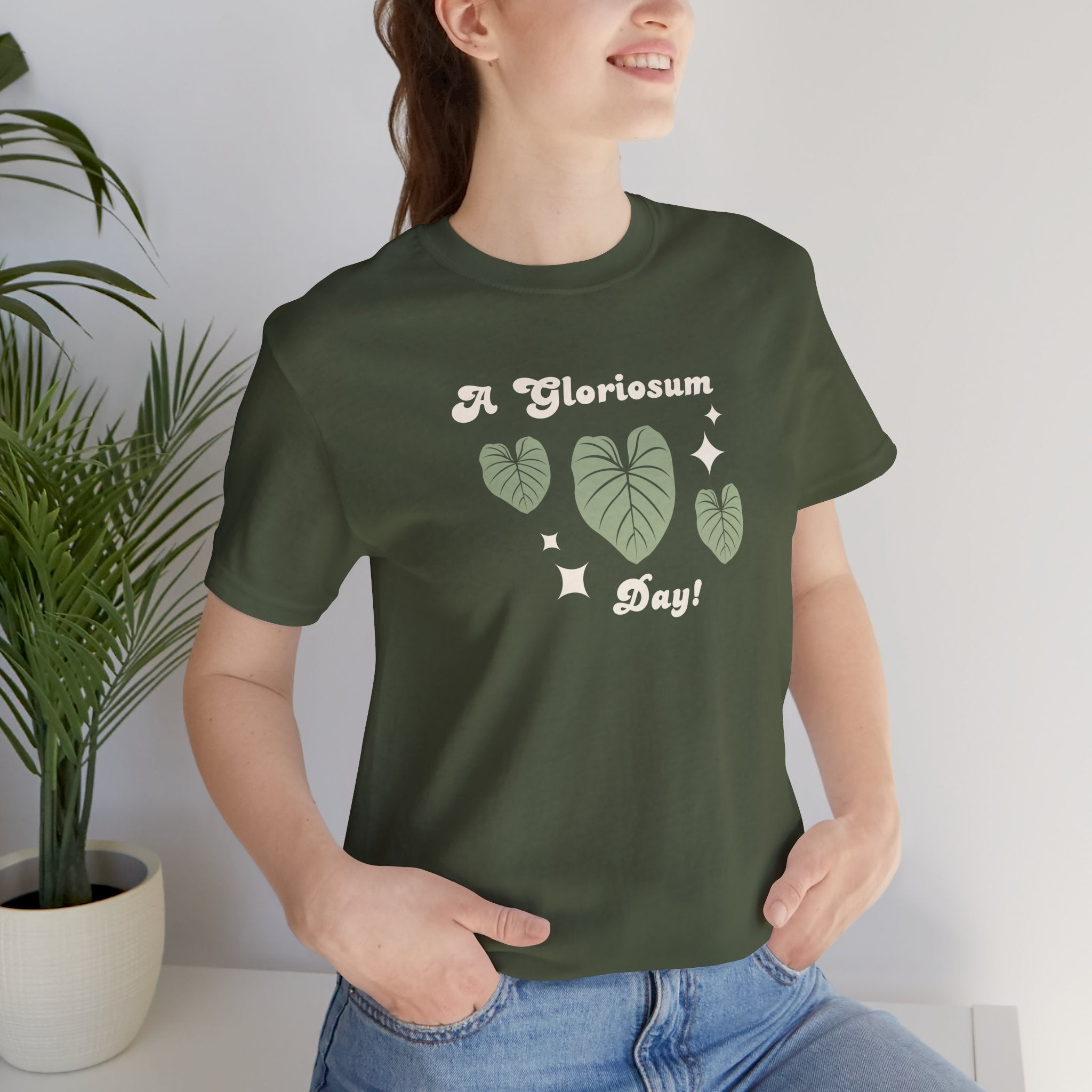 A woman wearing a military green t-shirt with text that says "A gloriosum day!"  The shirt design also has leaves from the philodendron gloriosum plant. 
