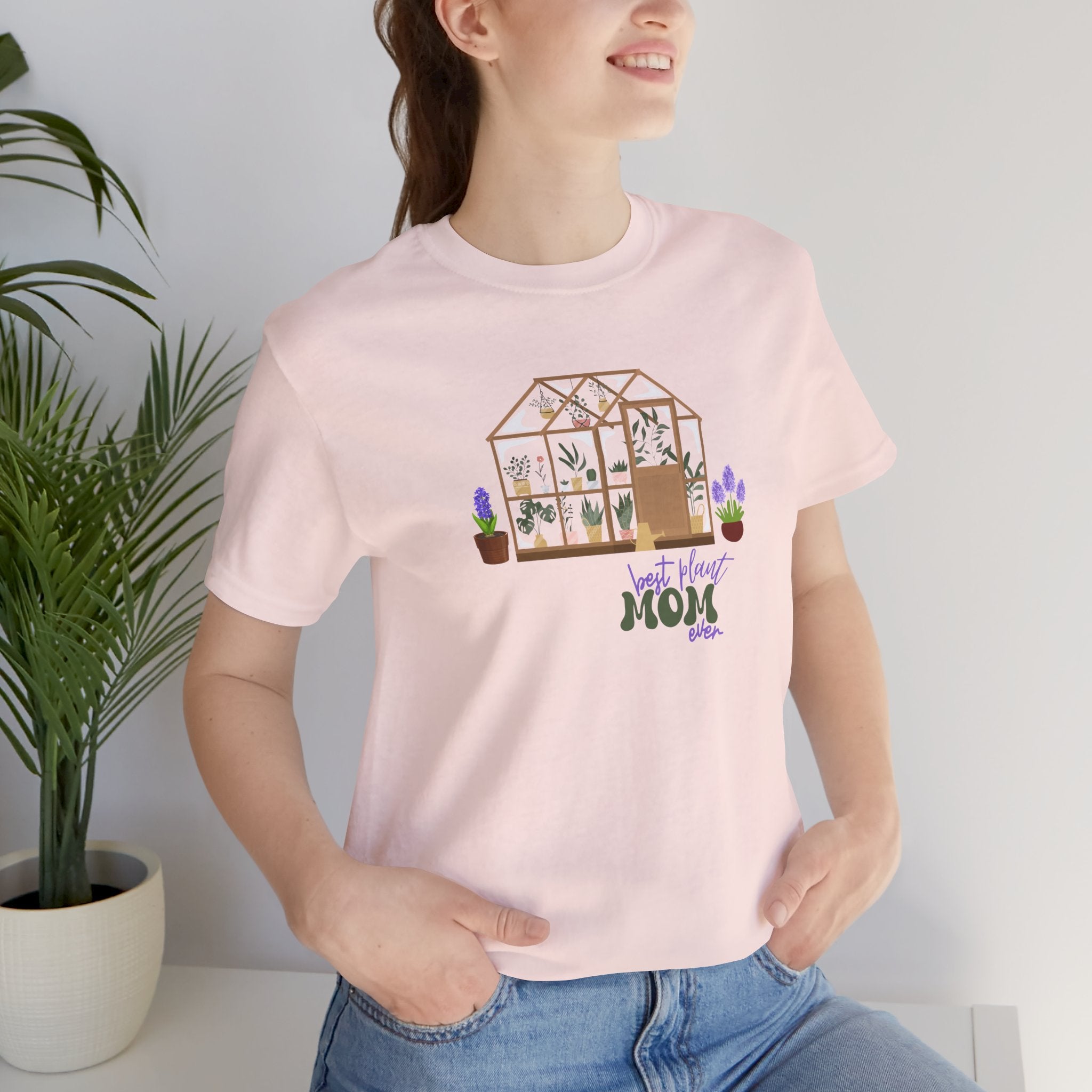 A lady wearing a jersey short sleeve tee in soft pink color with a greenhouse design.  There is also printed text that says best plant mom ever.