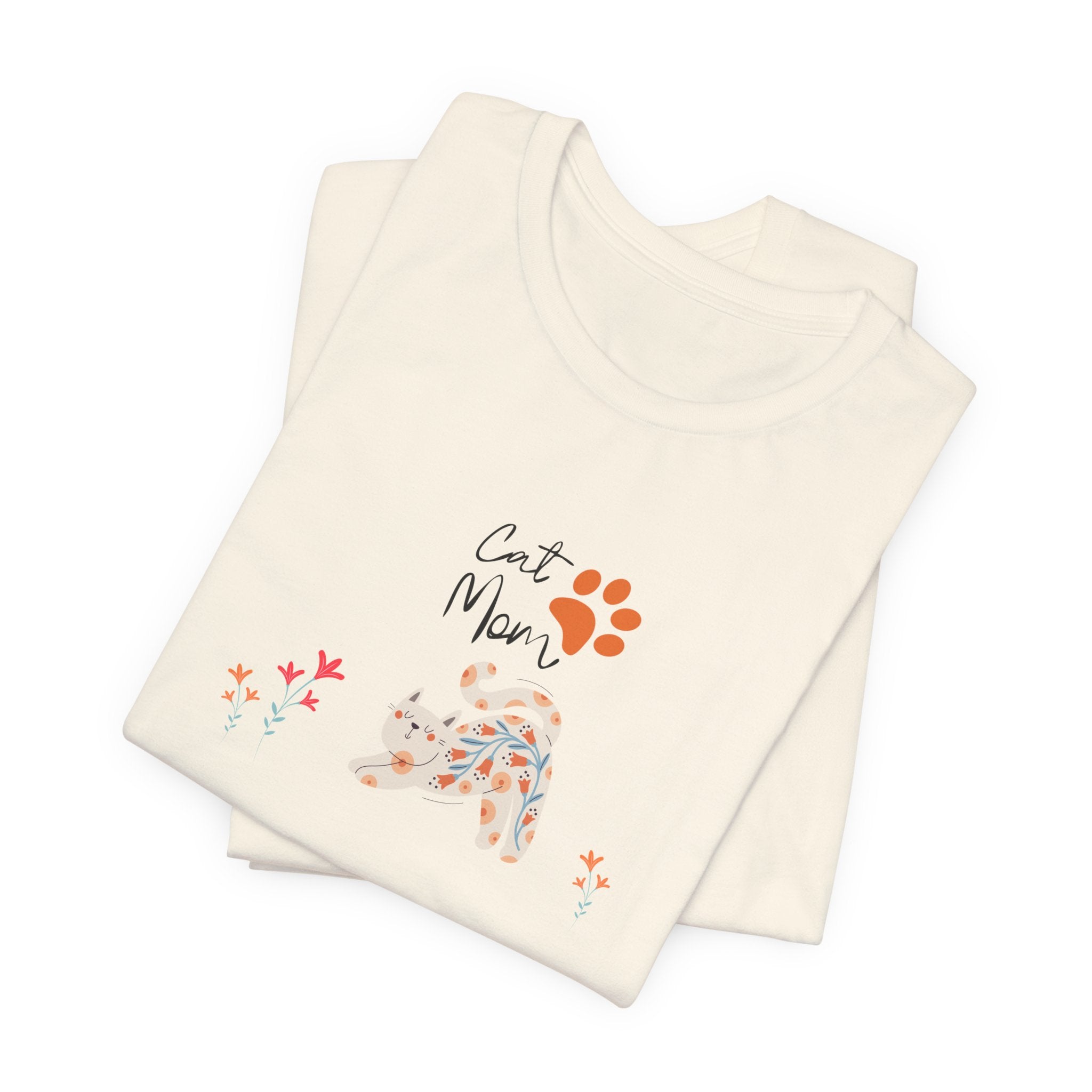A folded natural color t-shirt with the words "cat mom," a floral cat, and some flowers printed on the front of the shirt.