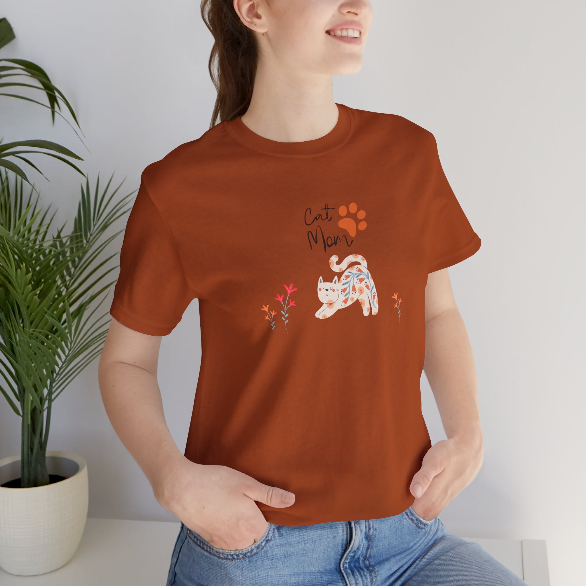 A woman wearing an autumn colored t-shirt with text that says "Cat Mom."  There is also a cat print, a floral cat, and flowers around the cat design. 