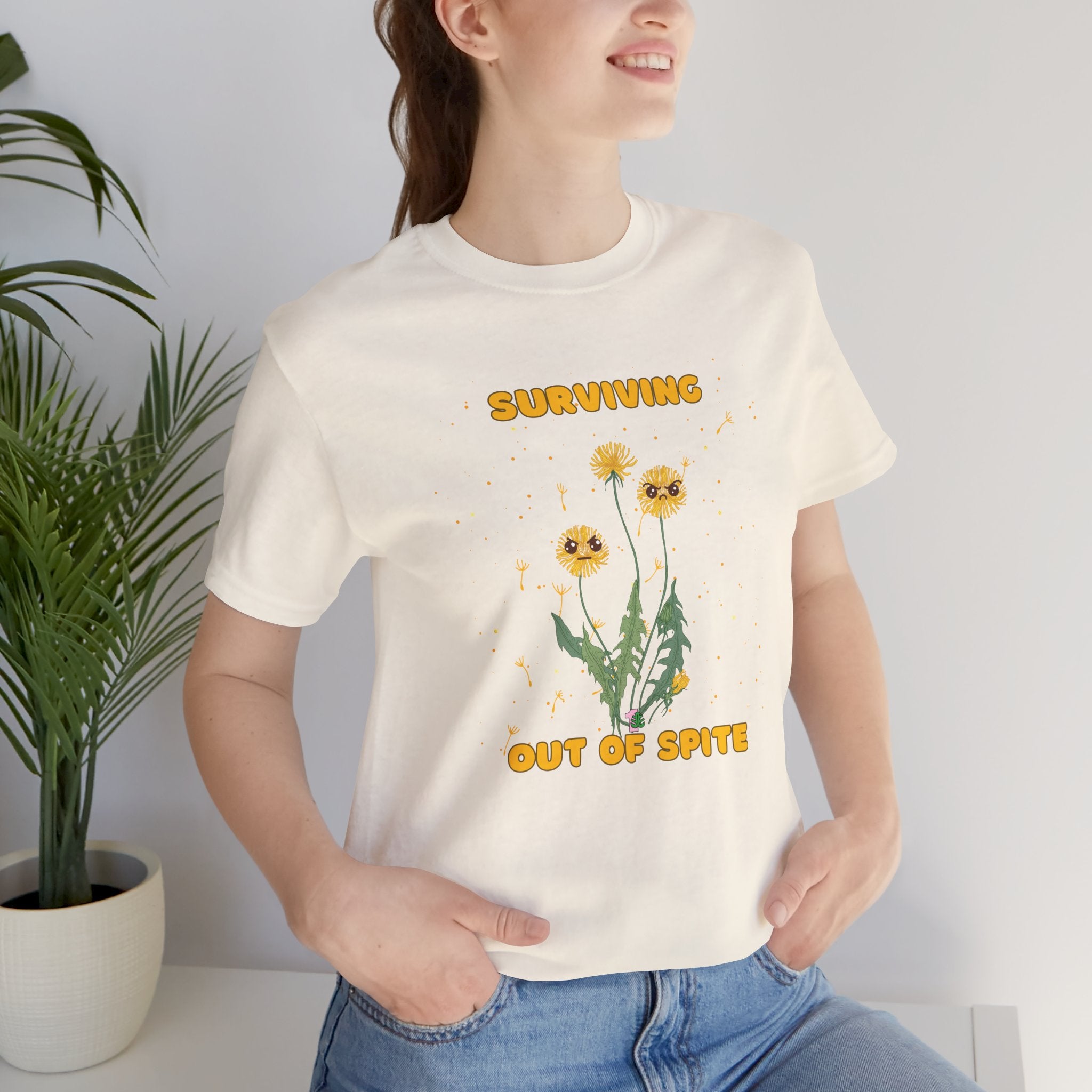 A person wearing a natural color t-shirt with yellow "surviving out of spite" funny text and dandelion plants with a funny look printed on the front of the shirt. 