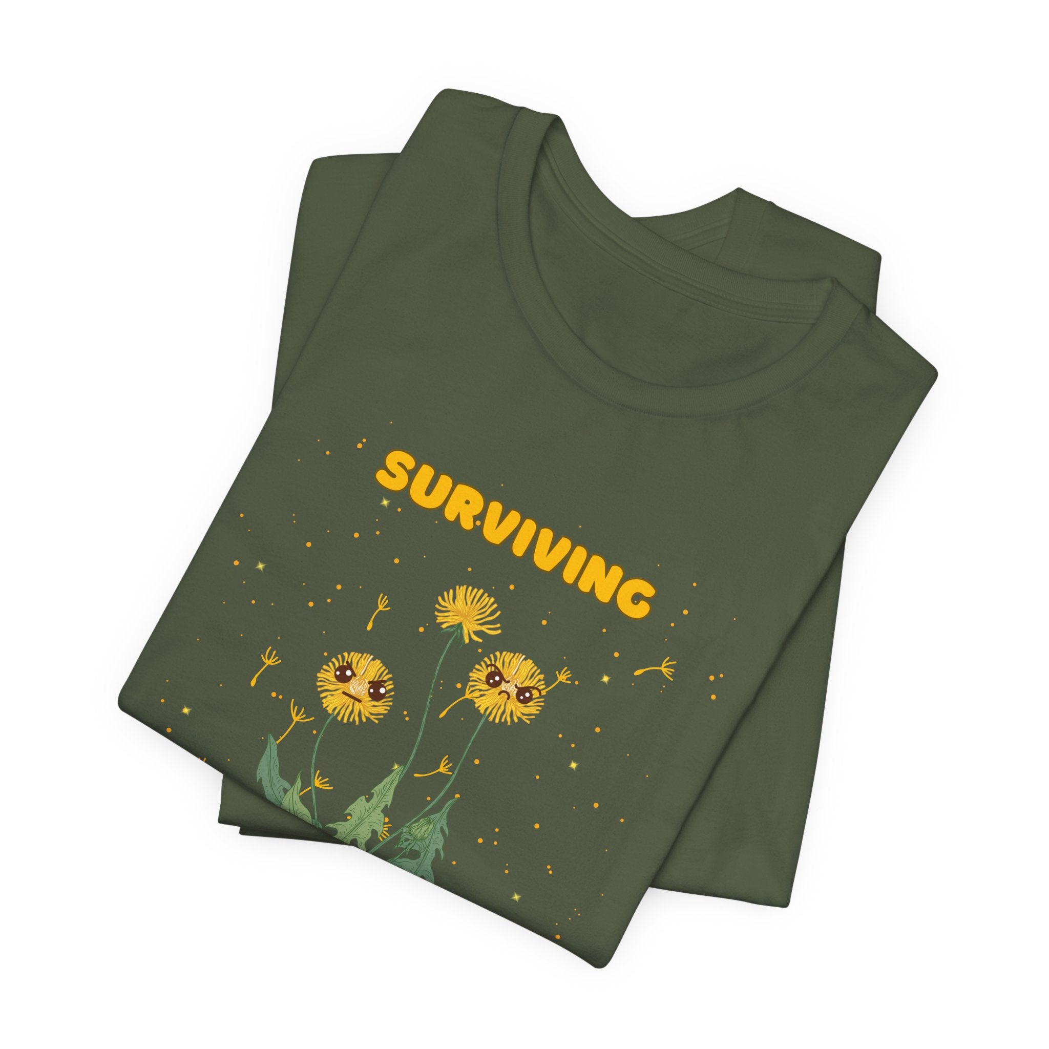A folded military green color t-shirt with yellow and green "surviving out of spite" funny text.  There is also dandelion plants with a funny look printed on the front of the shirt. 