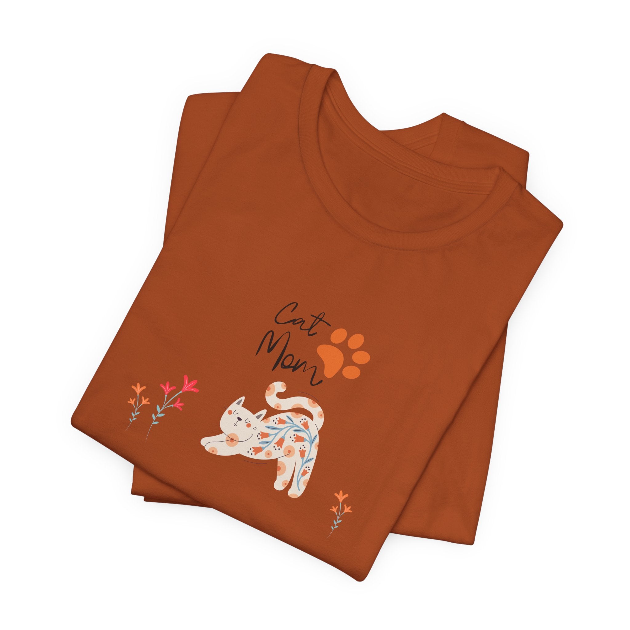 A folded autumn colored t-shirt with text that says "Cat Mom."  There is also a cat print, a floral cat, and flowers around the cat design. 