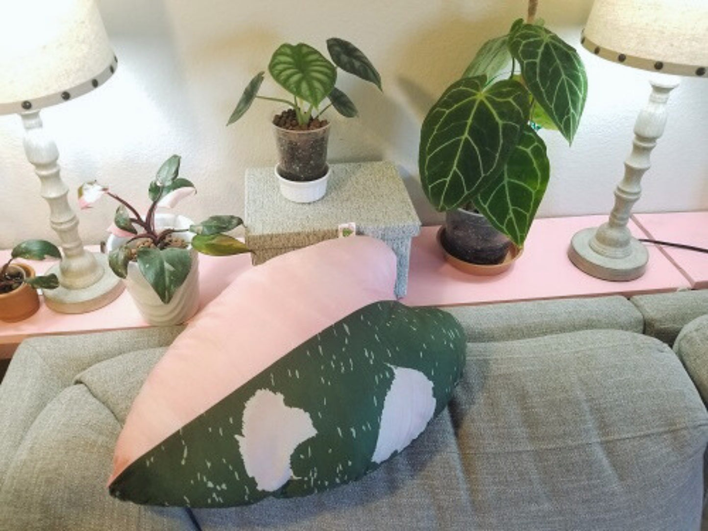 A philodendron pink princess heart-shaped throw pillow. The pillow has a variegated design with one half of the pillow being pink and the other half being green.  The pillow is a replica of a leaf print based on a natural philodendron pink princess plant leaf.  In this picture, the pillow is displayed on a gray couch, surrounded by various houseplants. The pillow is reversible, soft, velvety, and plush. It is the perfect gift ideal for plant lovers. Perfect for sofas, beds, and chairs.