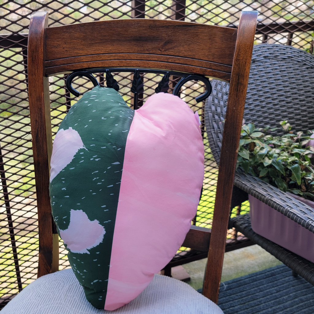 A decorative plant pillow on a wooden chair outdoors.  A leaf shaped pillow with a variegated pattern that resembles the pattern on a philodendron pink princess plant. 