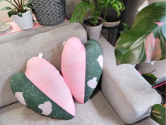Two philodendron pink princess heart and leaf shaped throw pillows on a sofa. The pillows have a variegated design with one half of the pillow being pink and the other half being green.  Each pillow is a replica of a leaf print based on a natural philodendron pink princess plant leaf.  In this picture, the pillows are displayed on a gray couch, surrounded by various houseplants. The pillows are soft, velvety, and plush. They are the perfect gift ideal for plant lovers and house plant collectors.