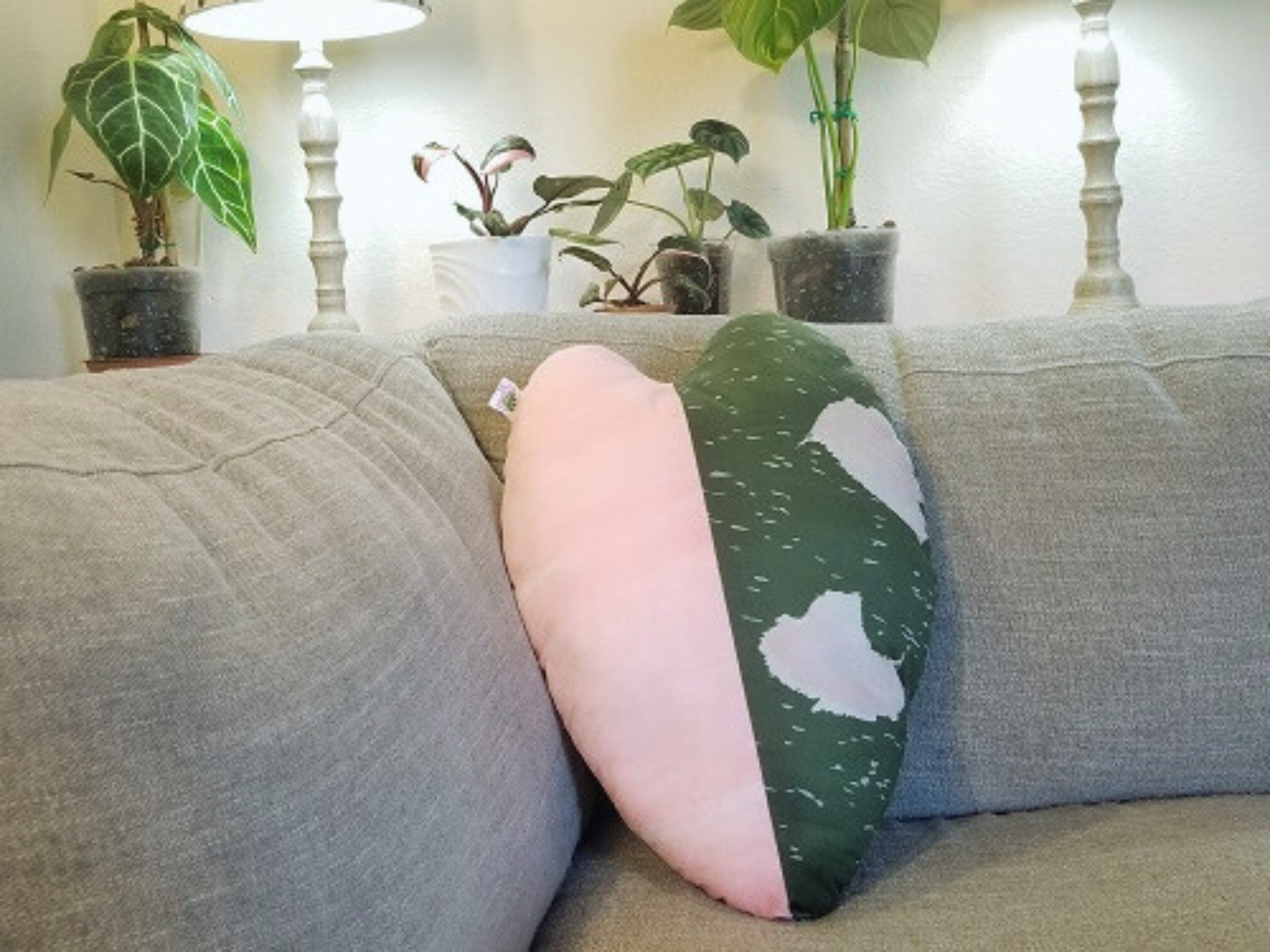 A philodendron pink princess throw pillow that is heart and leaf shaped. The pillow has a variegated design with one half of the pillow being pink and the other half being green.  The pillow is a replica of a leaf print based on a natural philodendron pink princess plant leaf.  In this picture, the pillow is displayed on a gray couch, surrounded by various houseplants. The pillow is reversible, soft, velvety, and plush. It is the perfect gift ideal for plant lovers. Perfect for sofas, beds, and chairs.